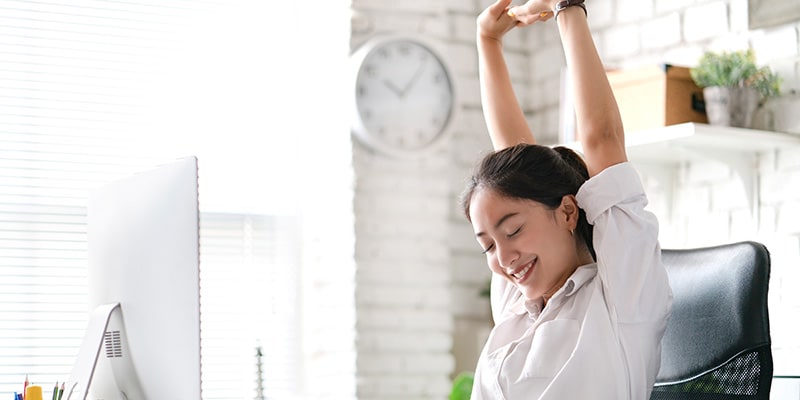A young Asian woman is stretching high with her arms whilst sat at her desk